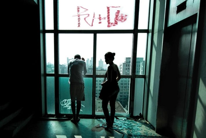 A still of a man and a woman standing in front of floor-to-ceiling windows with broken glass next to them.
