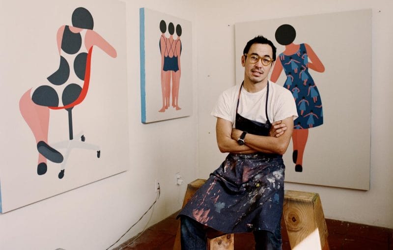A still of Geoff McFetridge in a paint-splattered apron seated in front of three of his artworks.