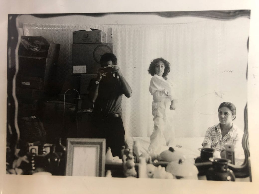An old photograph of a three people in a bedroom: a man holding a camera, a child, and a woman sitting.