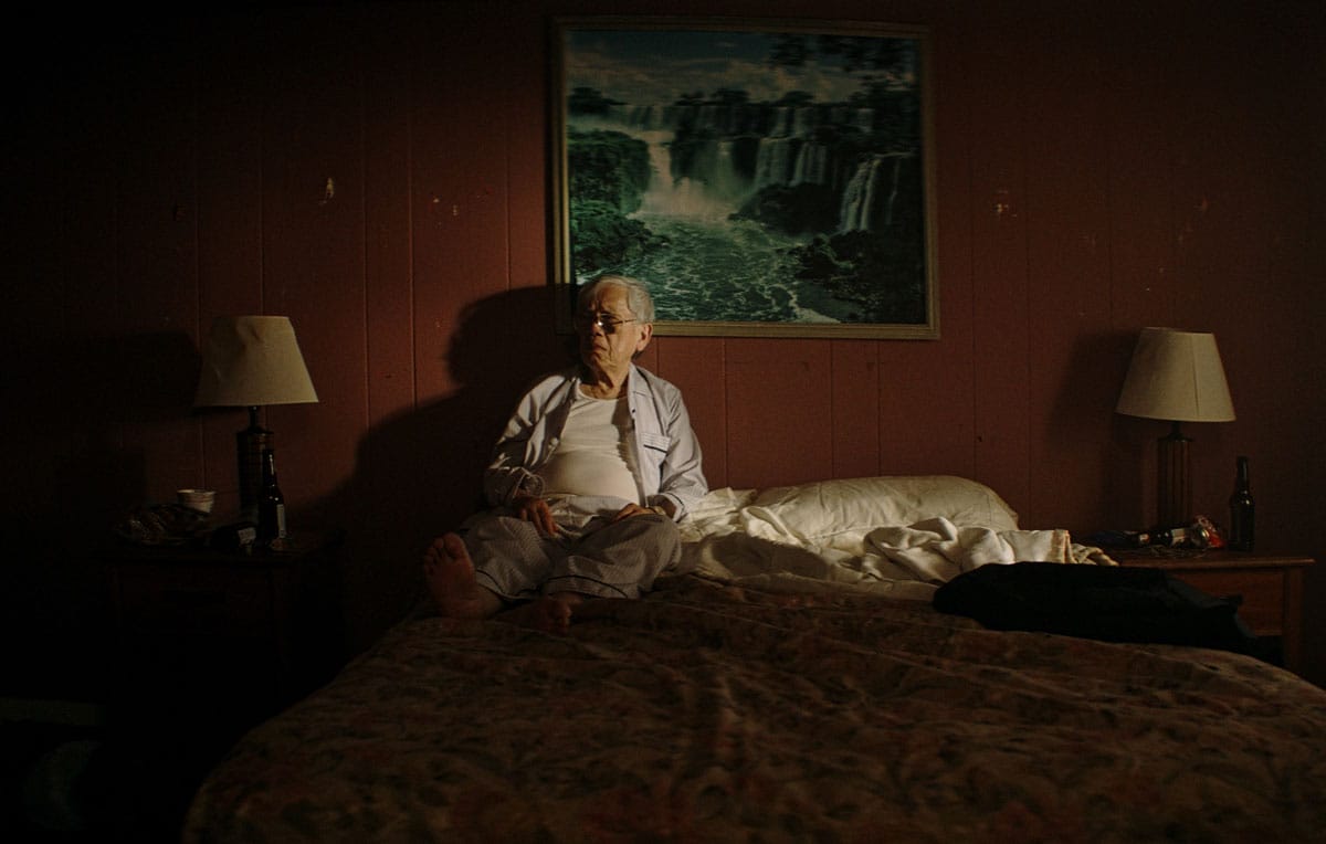 Long Má seated on a bed in a room with an orange wall with a picture of a waterfall.