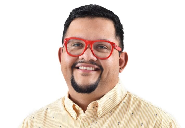 A portrait of Juan Carlos Noguera wearing thick-rimmed red glasses.