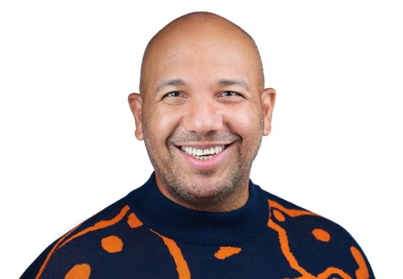 A portrait of Ramon Tejada smiling in a navy and orange sweater.