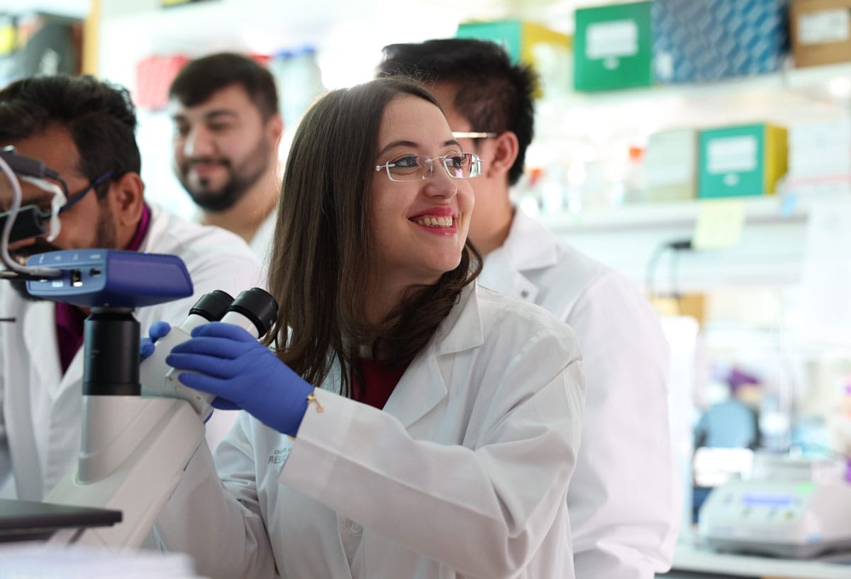 Gerta Hoxhaj smiles in front of a microscope with her colleagues in the background.