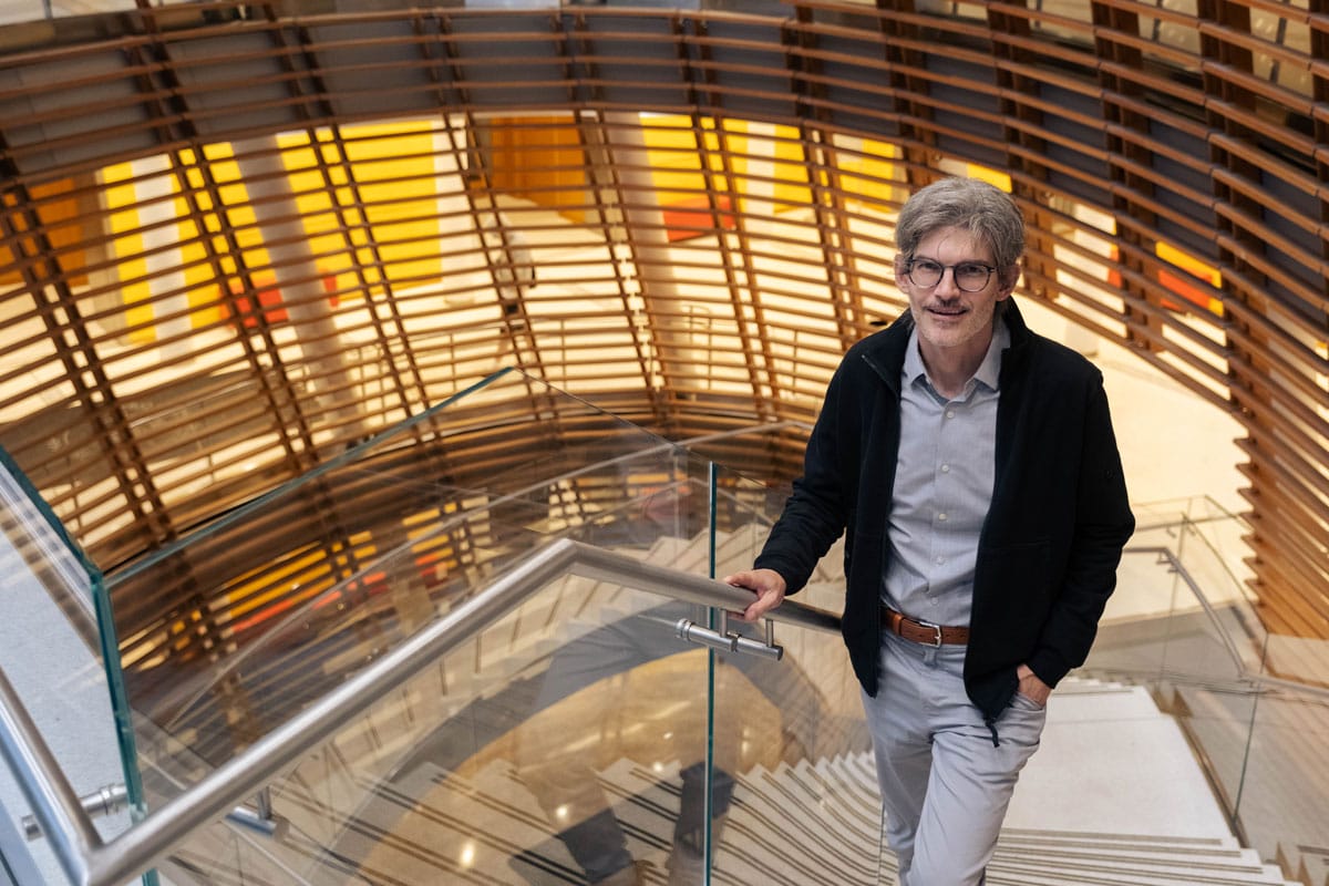 Luciano Marraffini standing on the stairs inside The Rockefeller University with wooden-screened railings behind him. 