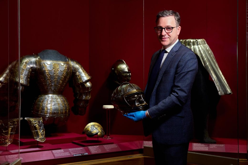 Pierre Terjanian holding a medieval helmet in front of an exhibit case of armor. 