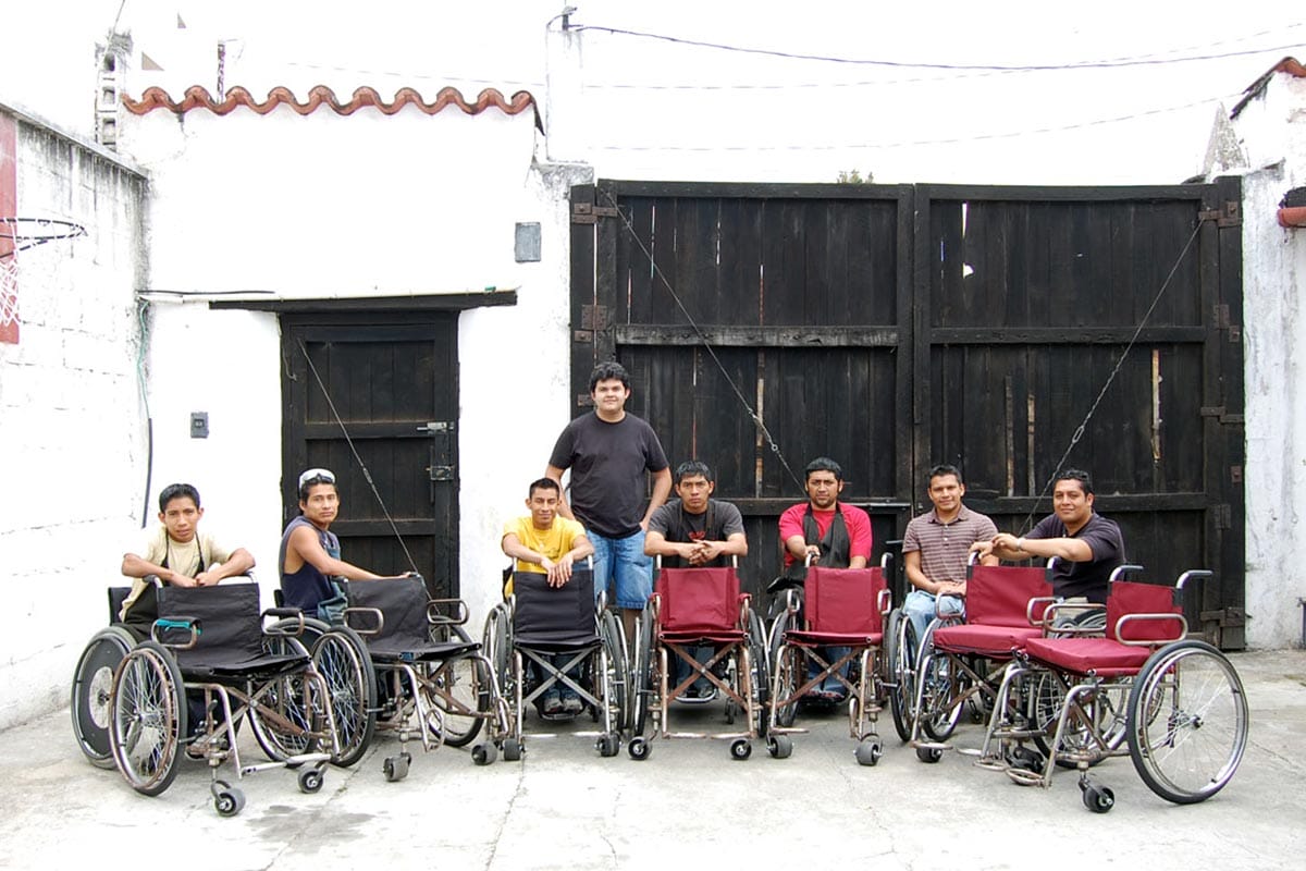 A young Juan Carlos Noguera smiles for a photo with a group of wheelchair-users in front of a black and white wall.