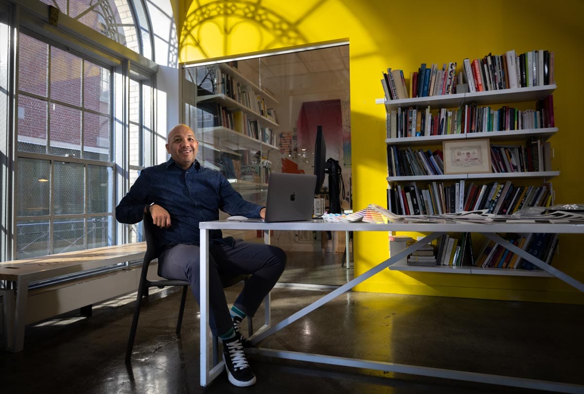 Ramon Tejada sitting at a desk with the window frame casting a shadow on the wall behind him.