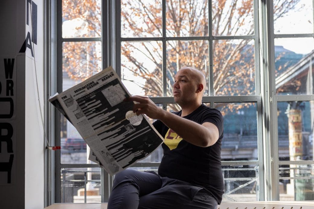 Ramon Tejada sitting in front of a large window reading a newspaper.