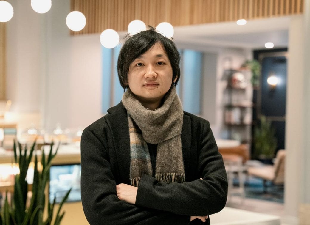 Takanori Takebe wearing a scarf and standing in a coffee shop with his arms crossed.