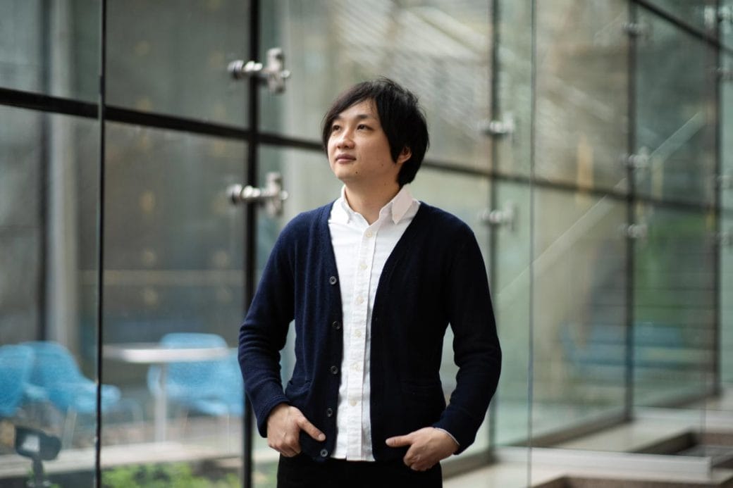 Takanori Takebe stands with his hands in his pockets in front of a wall of glass windows. 