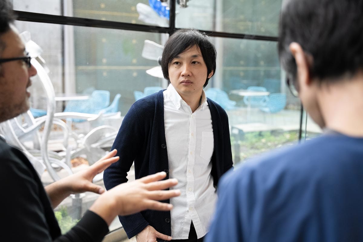Takanori Takebe talks with his colleagues in front of large, bright windows.