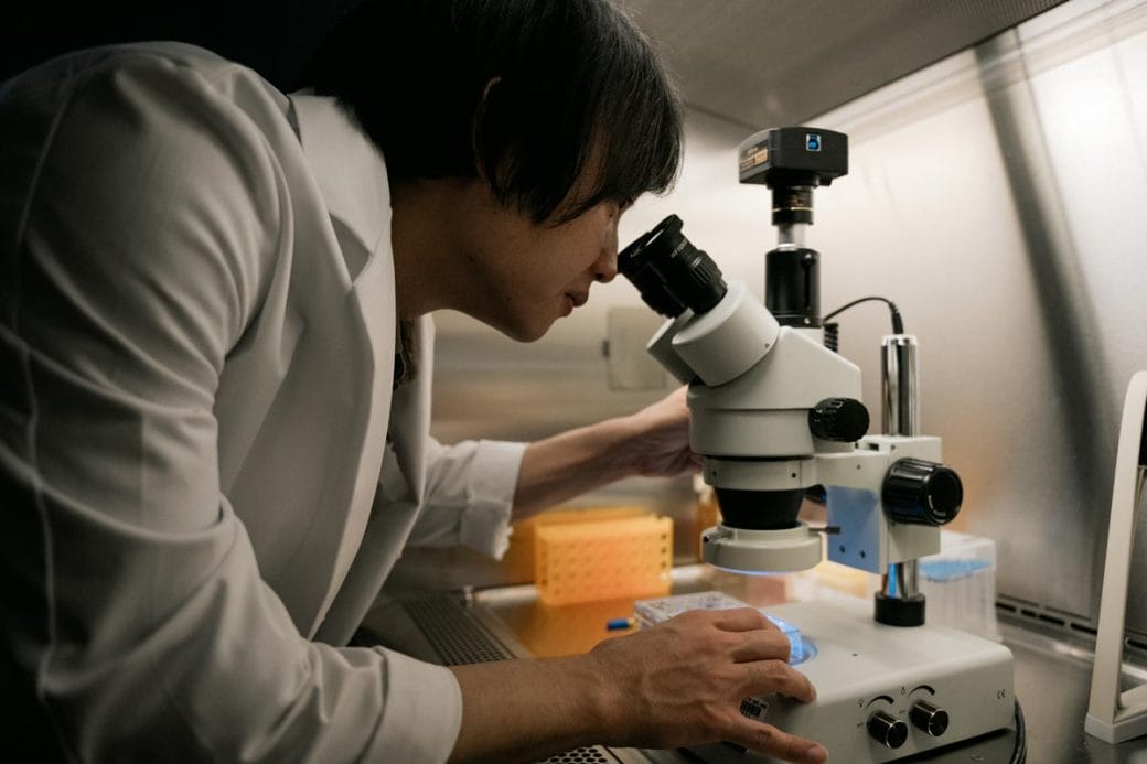 Takanori Takebe wearing a white lab coat and looking into a microscope.