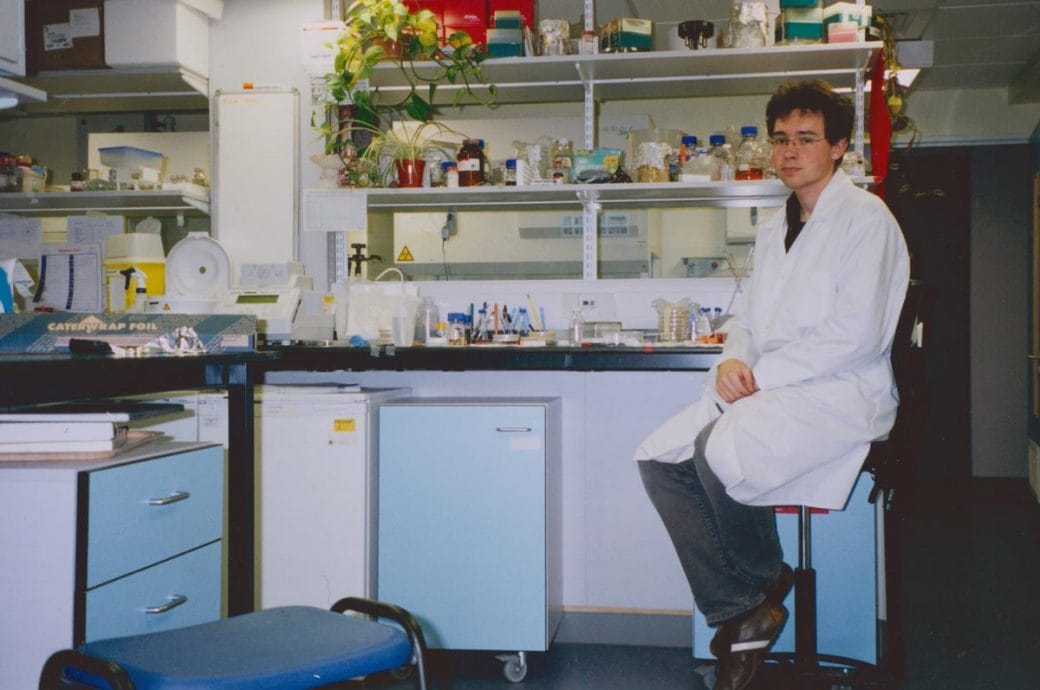 A young Tomasz Nowakowski wearing a lab coat and sitting on a chair in a lab.