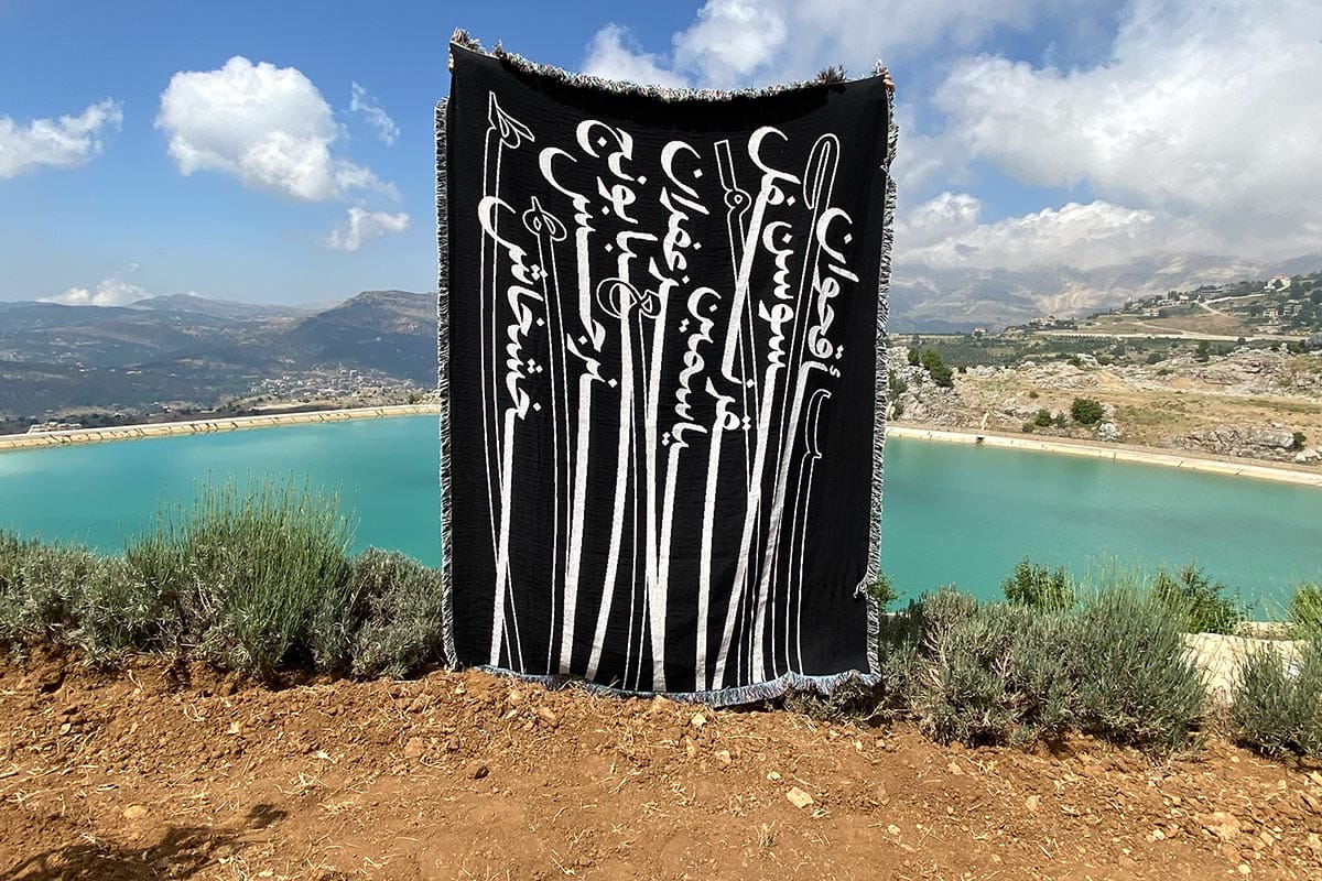 A large black tapestry with white Arabic typography in front of a serene view with crystal blue water.