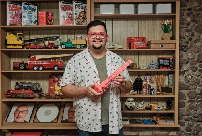 Juan Carlos Noguera standing in front of busy shelving holding a red string instrument that he designed.