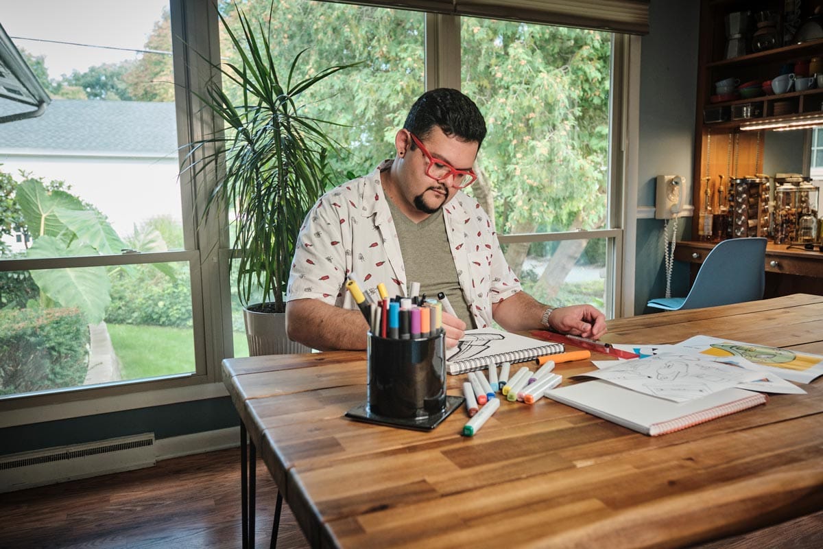 Juan Carlos Noguera sitting at a table drawing with colored markers with a lush garden showing through the large windows behind him. 