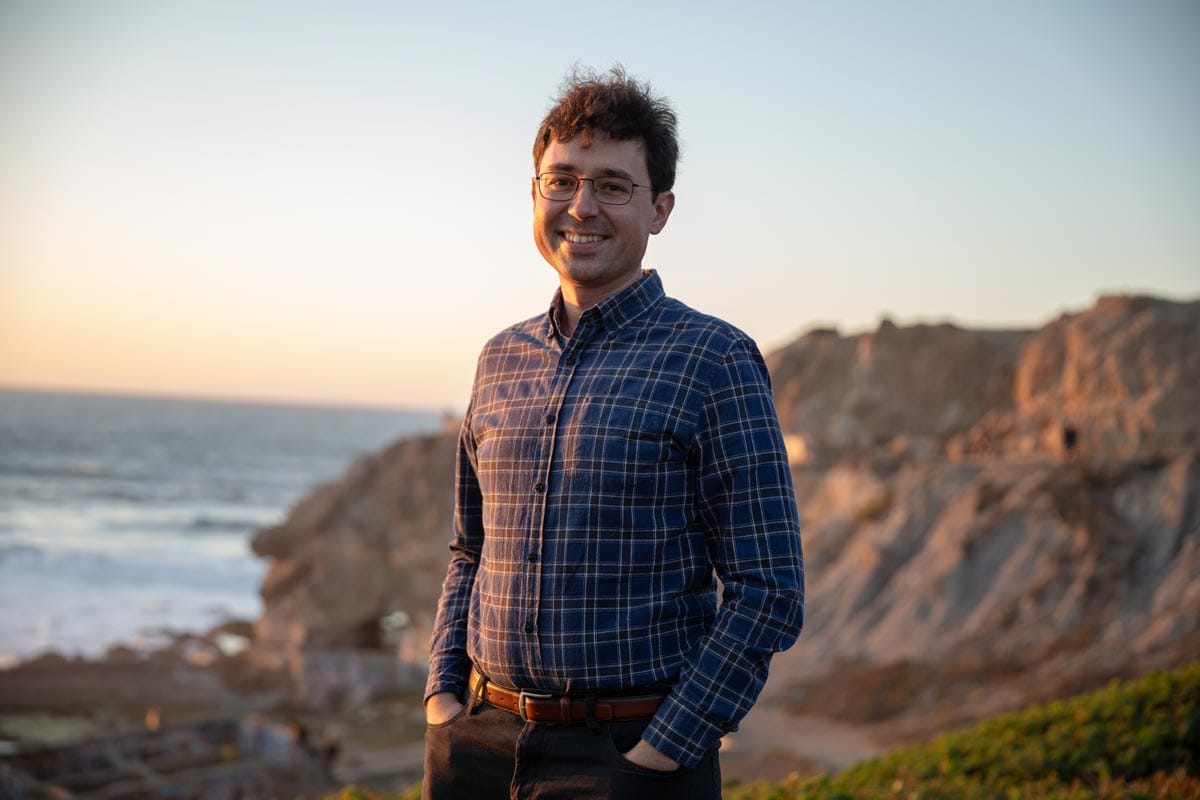 Tomasz Nowakowski standing on a beach with the ocean and rocky cliffs behind him. 