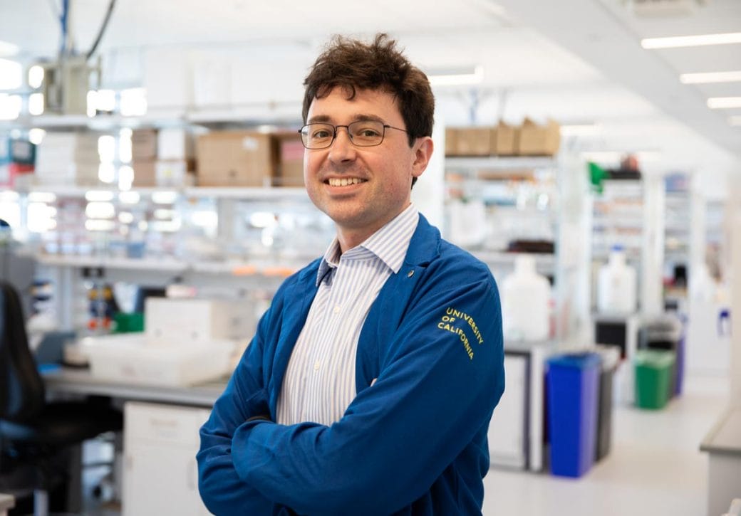 Tomasz Nowakowski, wearing a blue lab coat, stands in his lab smiling and folding his arms. 