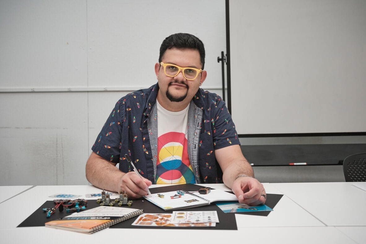 Juan Carlos Noguera sits working at a desk with books, designs, and small metal parts on it.