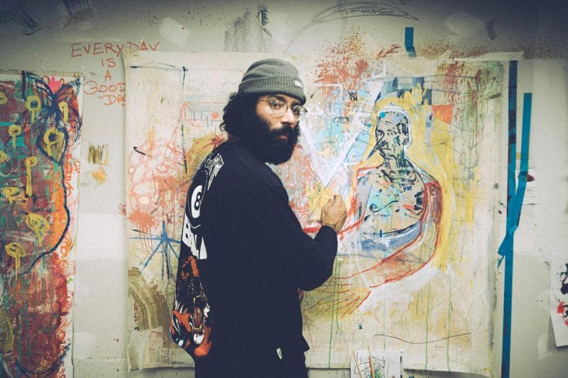 A man in a beanie and glasses standing in front of a multi-color artwork.