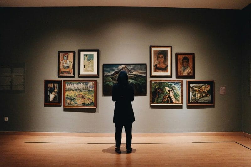A person standing in front of a gallery wall with art works hung on it.