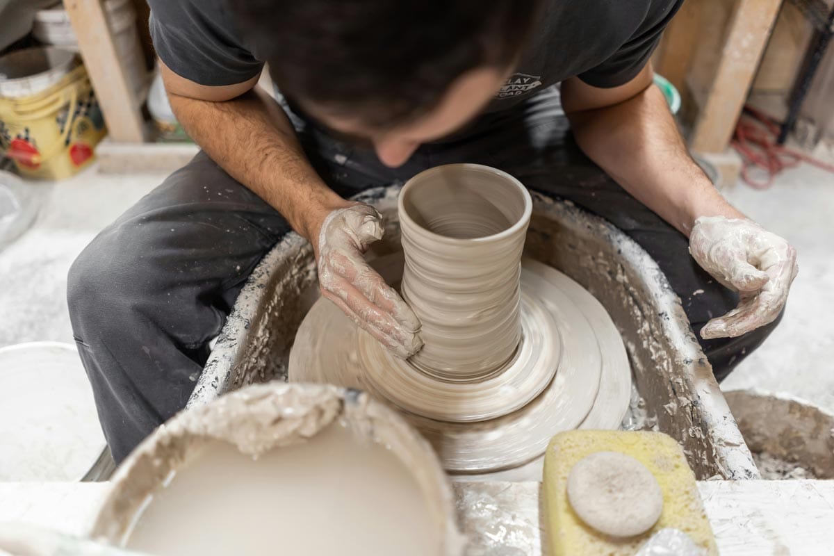 A man sits a pottery wheel shaping a cylindrical ceramic piece.