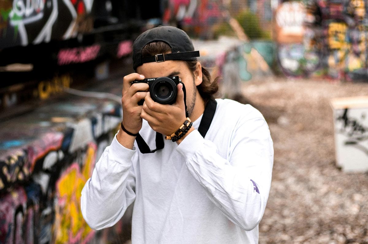 A man wearing a hat backwards taking a photo with his digital camera.