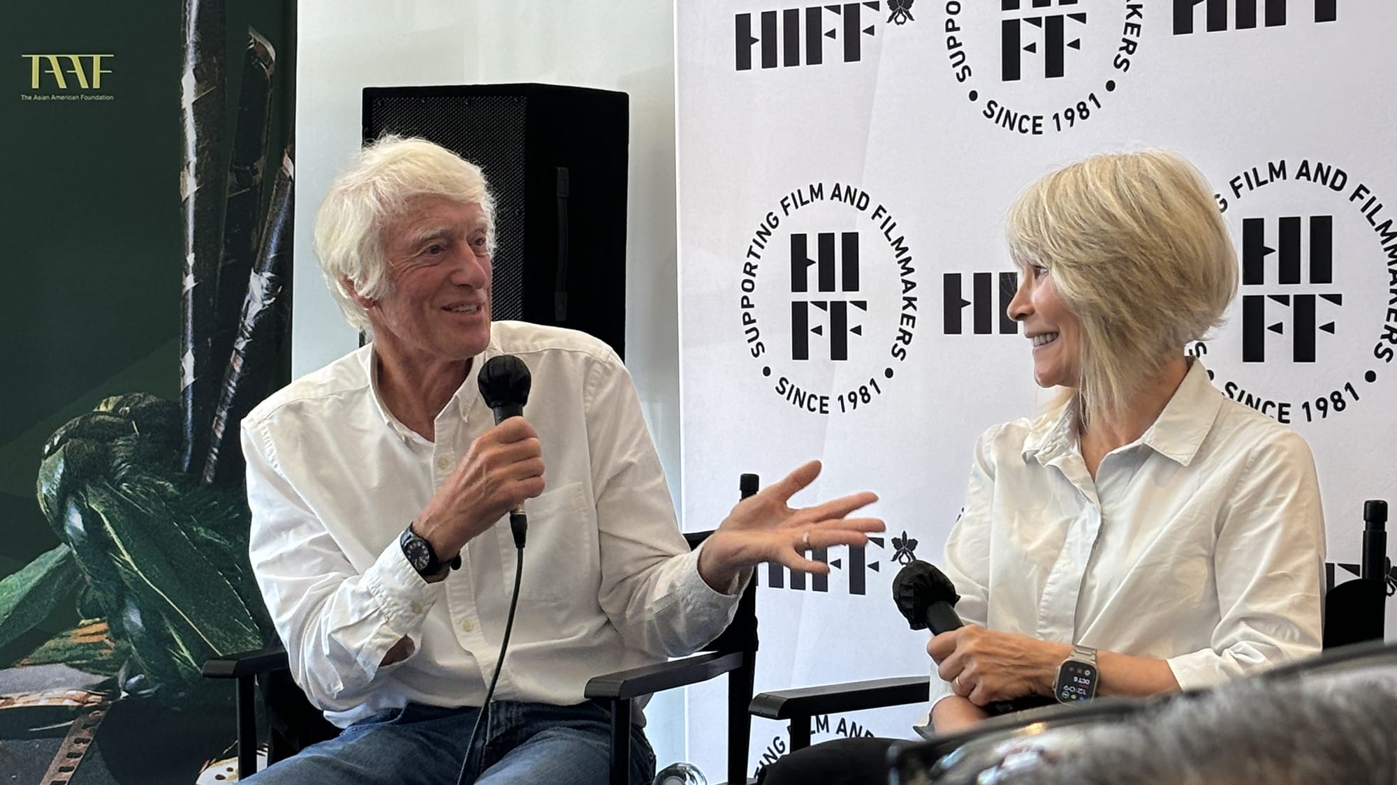 A man with white hair and a white shirt holds a microphone and gestures while looking at a woman with blonde hair, also holding a microphone, during a discussion.