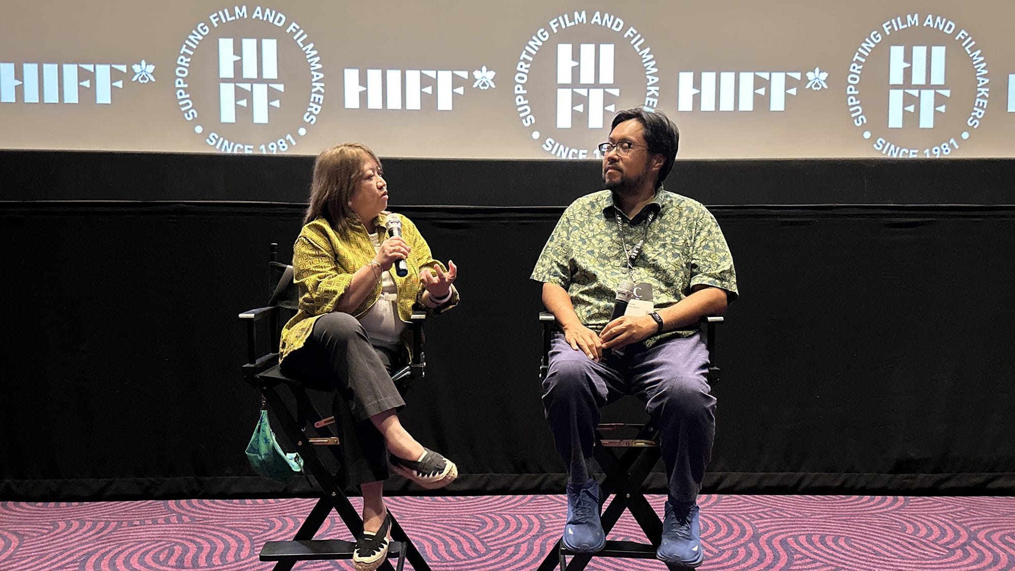 A woman sits in a chair holding a microphone and speaking to a man in a patterned shirt and glasses sitting in an adjacent chair.