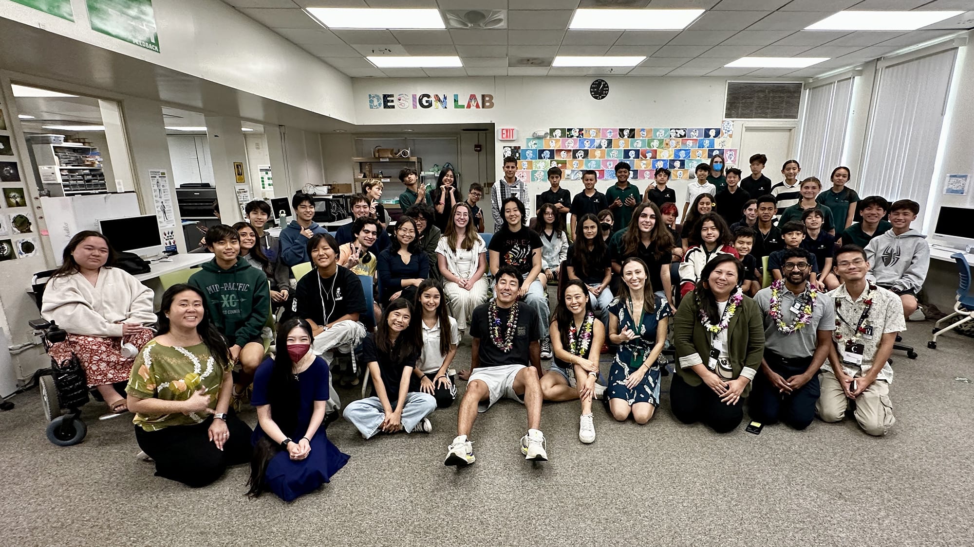 A large group of young people and a few adults, some wearing leis, pose for a photo in a classroom with DESIGN LAB written on the wall.