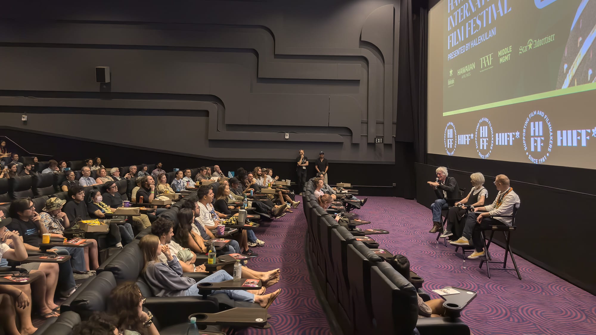 An audience fills a movie theater, with four people sitting on stools on stage at the front, in front of a large screen displaying HIFF logos.