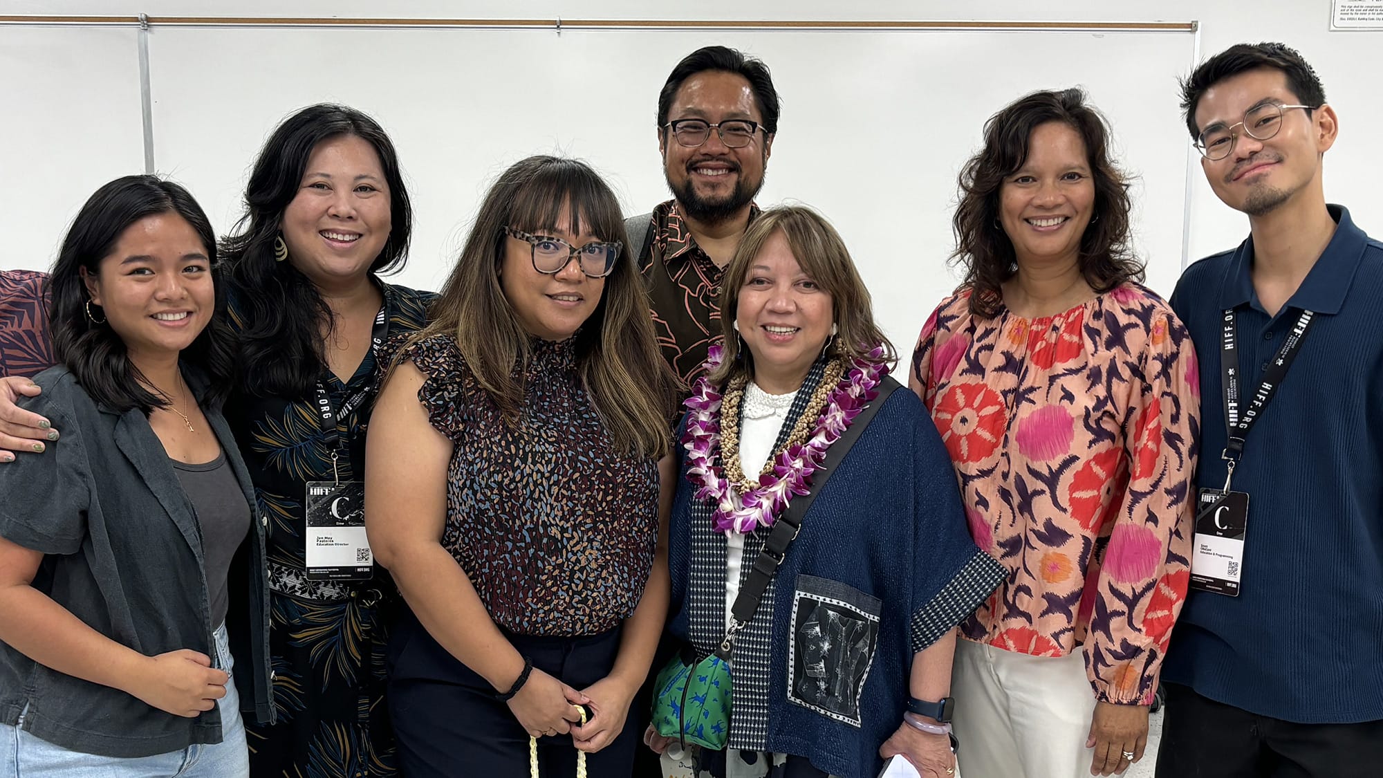 A group of six adults stands together, smiling at the camera, with one woman in the center wearing a lei.