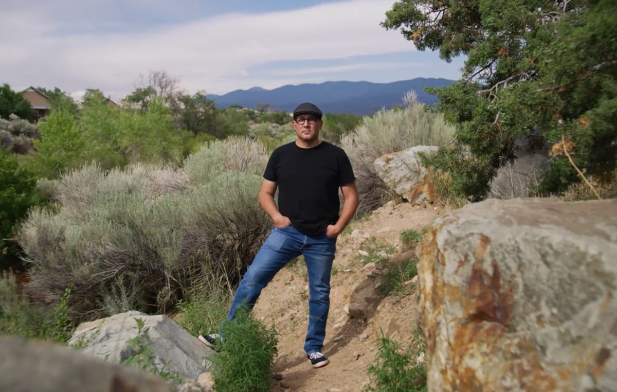 A full length portrait of Sergio Tapia standing on a river bank next to the Santa Fe River.