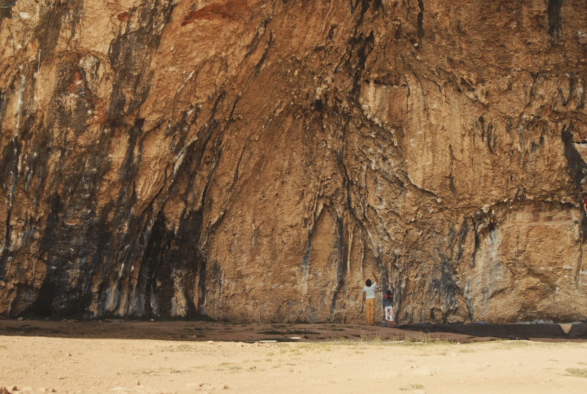 Rock climbers Ashima Shiraishi and Chris Sharma stand at the base of a rugged rock climb in Spain. The figures are dwarfed by the size of the landscape.