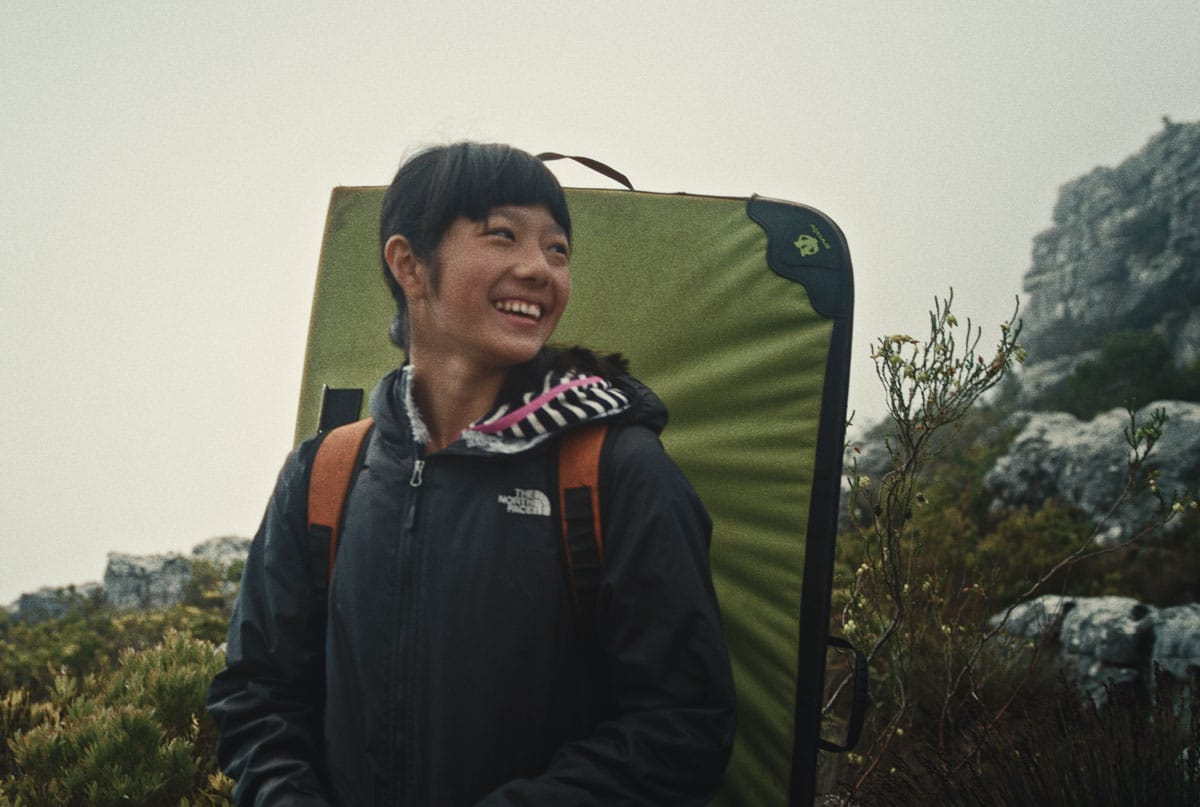 Ashima Shiraishi stands amid brush in Rocklands, South Africa. She is wearing a t-shirt and a windbreaker, and is carrying a bouldering crashpad on her back like a backpack.