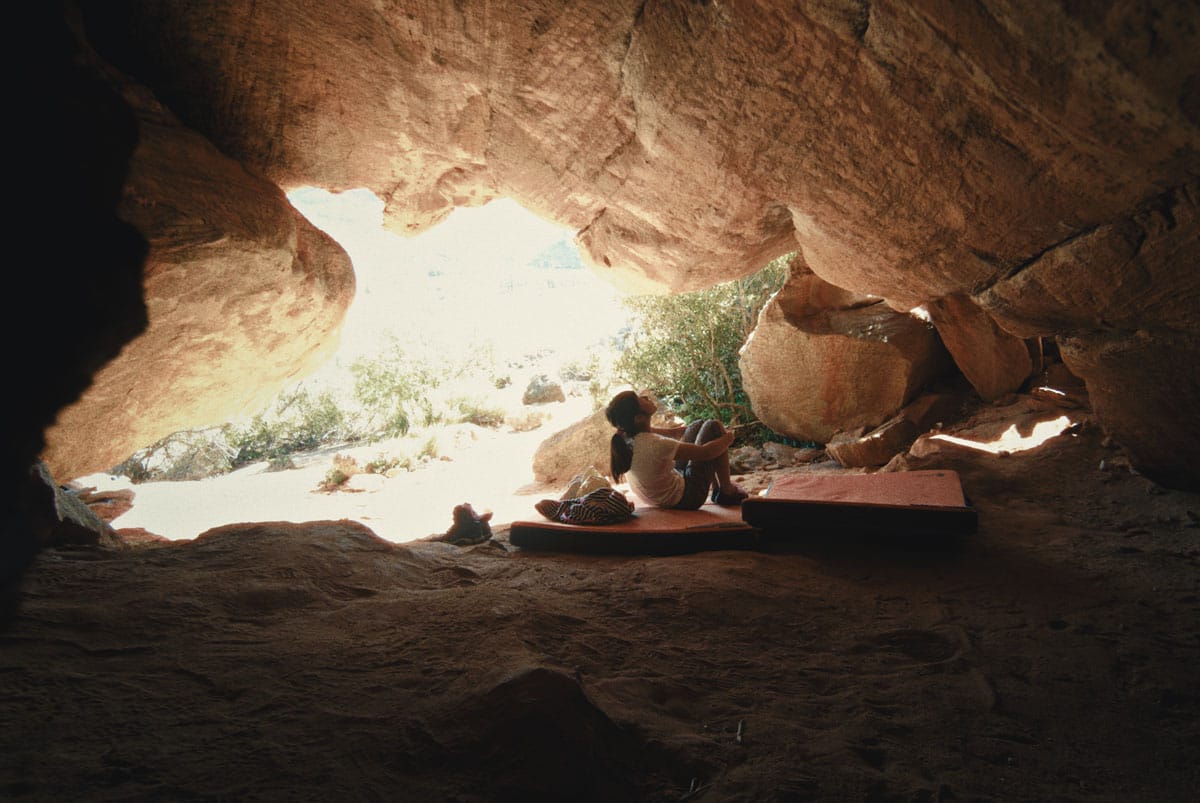 Ashima Shiraishi sits on the ground beneath a rocky arch overhead. She gazes up at the rock, studying a climbing route that she will attempt.