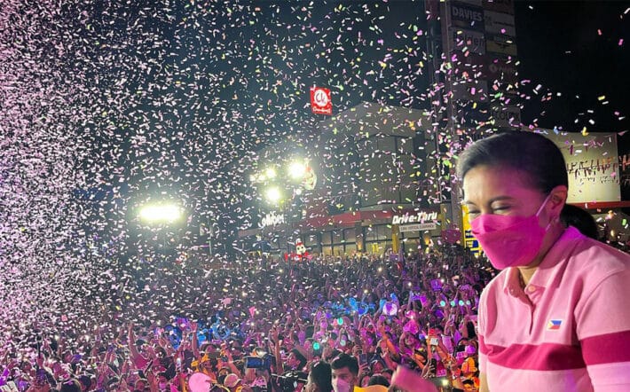 Former Vice President of the Philippines Leni Robredo wears a pink polo shirt embroidered with the flag of the Philippines. She stands on a stage amid a joyous crowd being showered with pink confetti during a rally during her run for President of the Philippines in 2022.