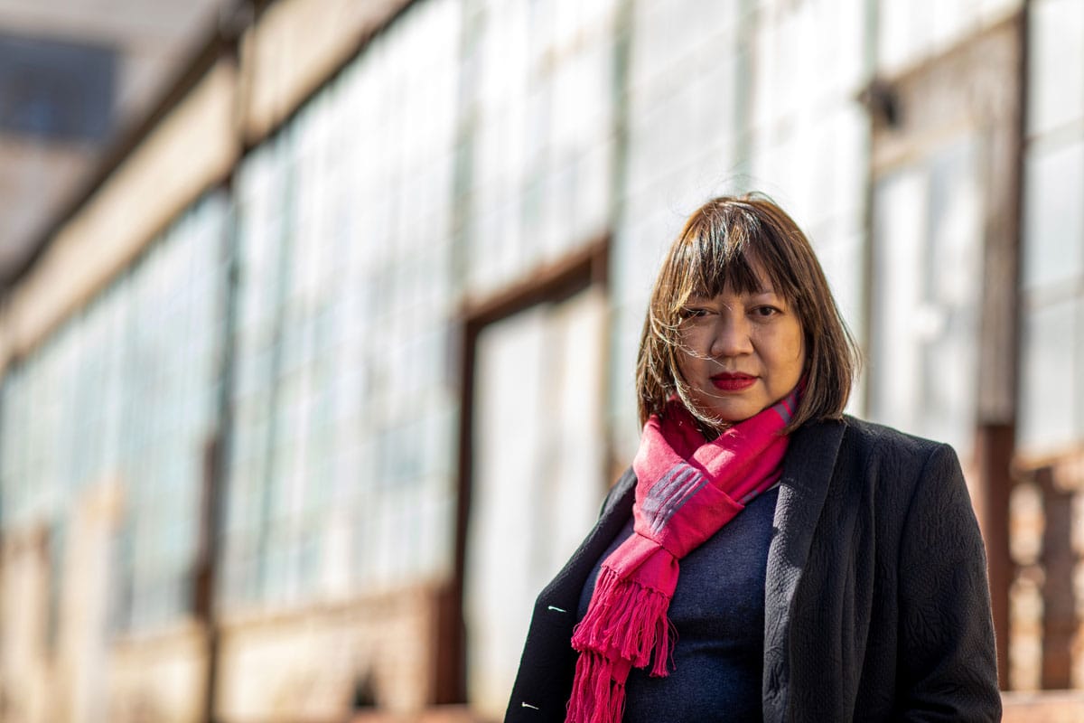 Ramona S. Diaz stands in front of a building in bright sunlight. She has chin-length hair styled in a bob, and is wearing a black jacket and a bright pink scarf.