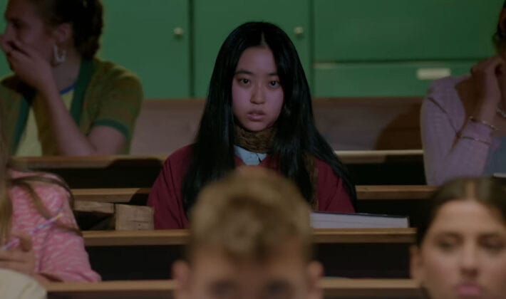 Wei sits in a classroom among students. She has long, black hair and her focus is locked-in in front of her.