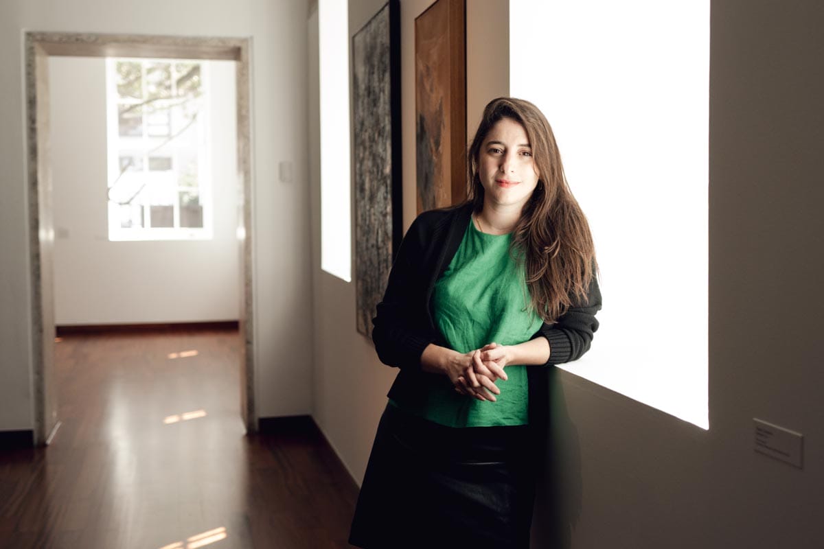 Aimé leans against a gallery wall with two large artworks visible behind her.