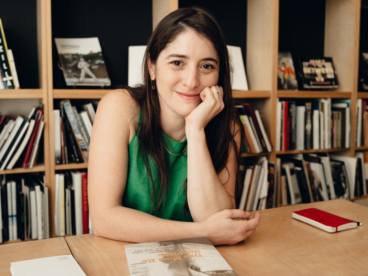 Aimé seated at a table with a bookcase behind her. She is resting her cheek on her hand.