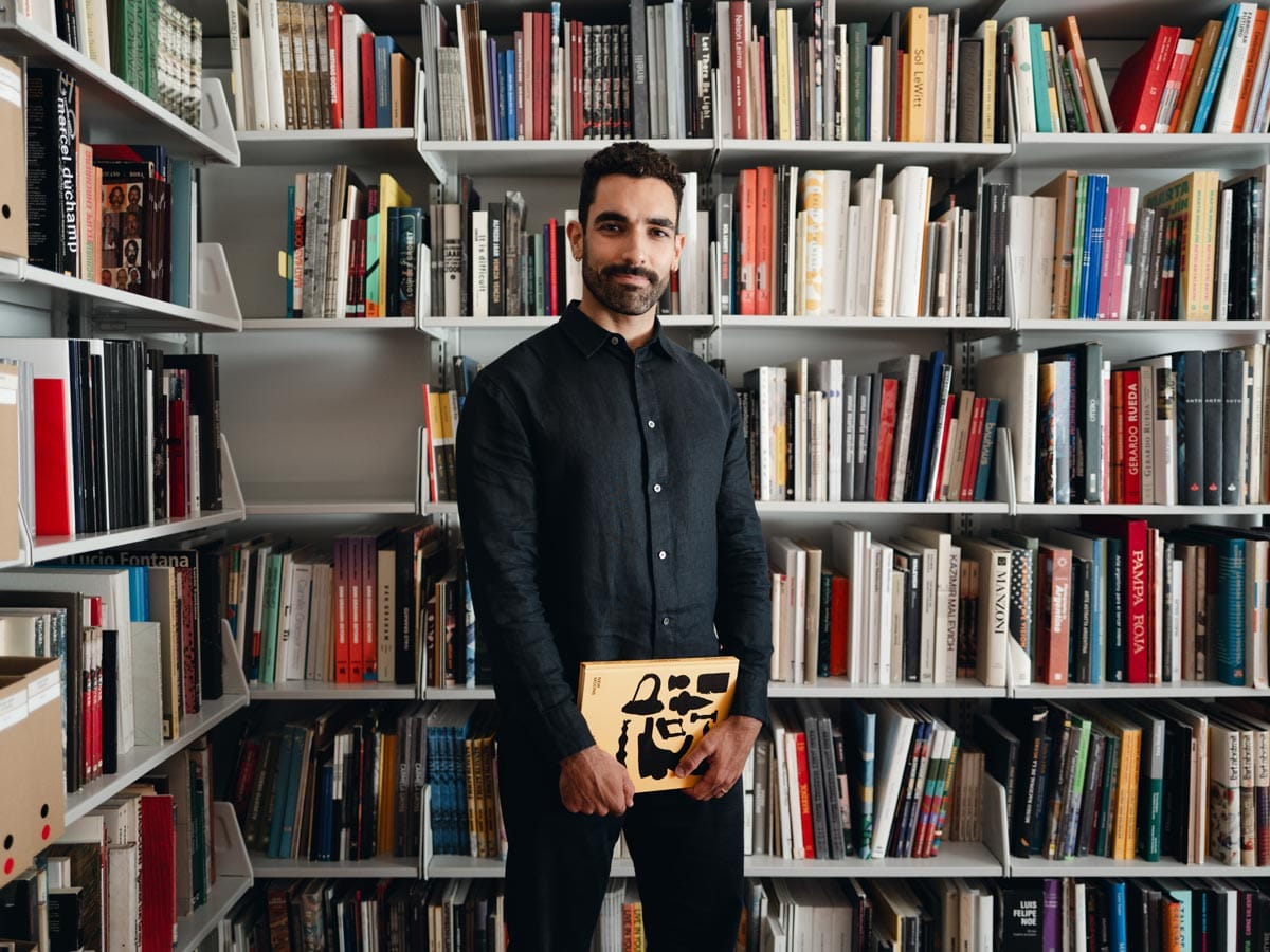 Bernardo holds a book whilst standing in front of a large bookcase filled with books.