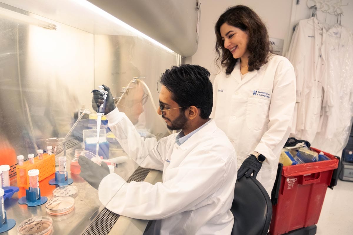 Elham observing her colleague conducting an experiment using a pipette in the lab.