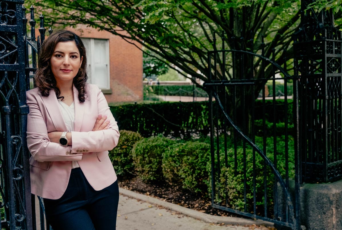 Elham leans against a wrought iron fence while wearing a pink blazer.