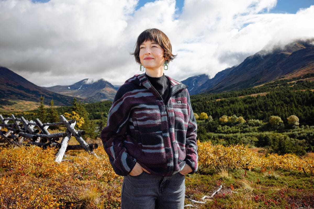 Francesca standing in nature in Alaska: there is mountain and brush behind her.