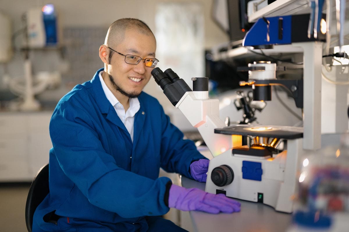 Guosong sits in front of a microscope wearing a blue lab coat.