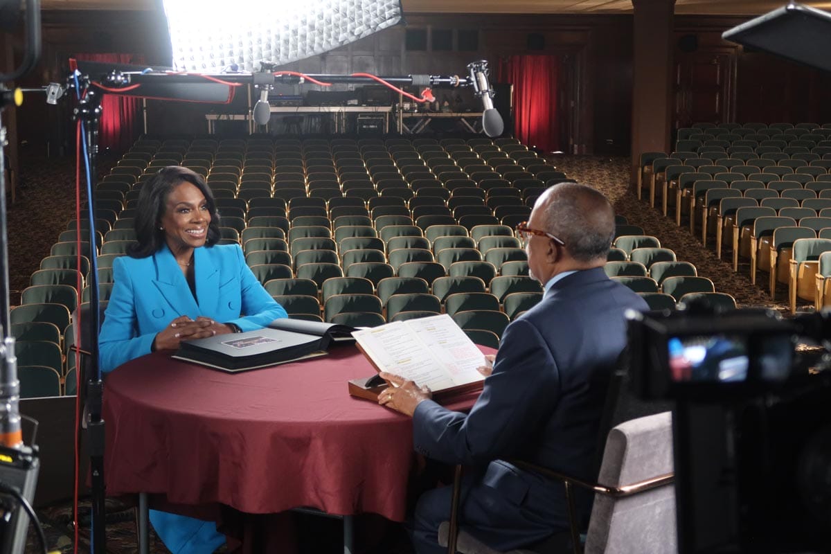 Henry Louis Gates Jr. and Sheryl Lee Ralph seated at a table on the set of Henry's PBS show: there is sound equipment and a bright light in the background.