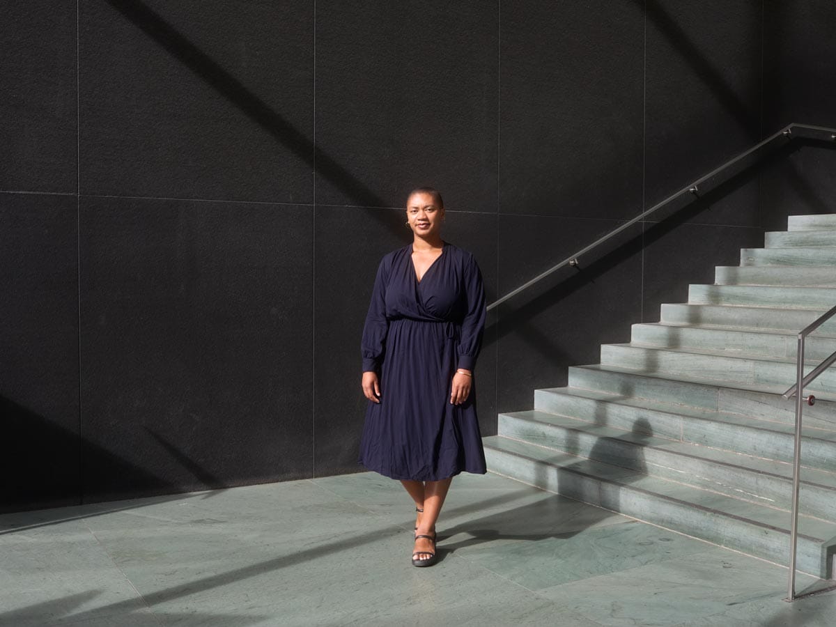 Oluremi wearing a navy dress standing at the bottom of a staircase in front of a stark black wall.