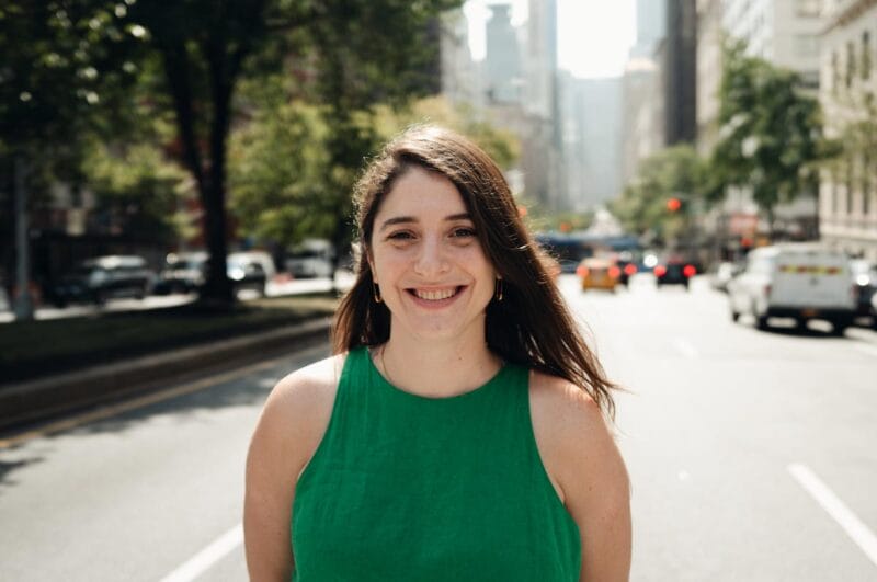 Aimé standing in a New York City crosswalk, smiling with city traffic behind her.