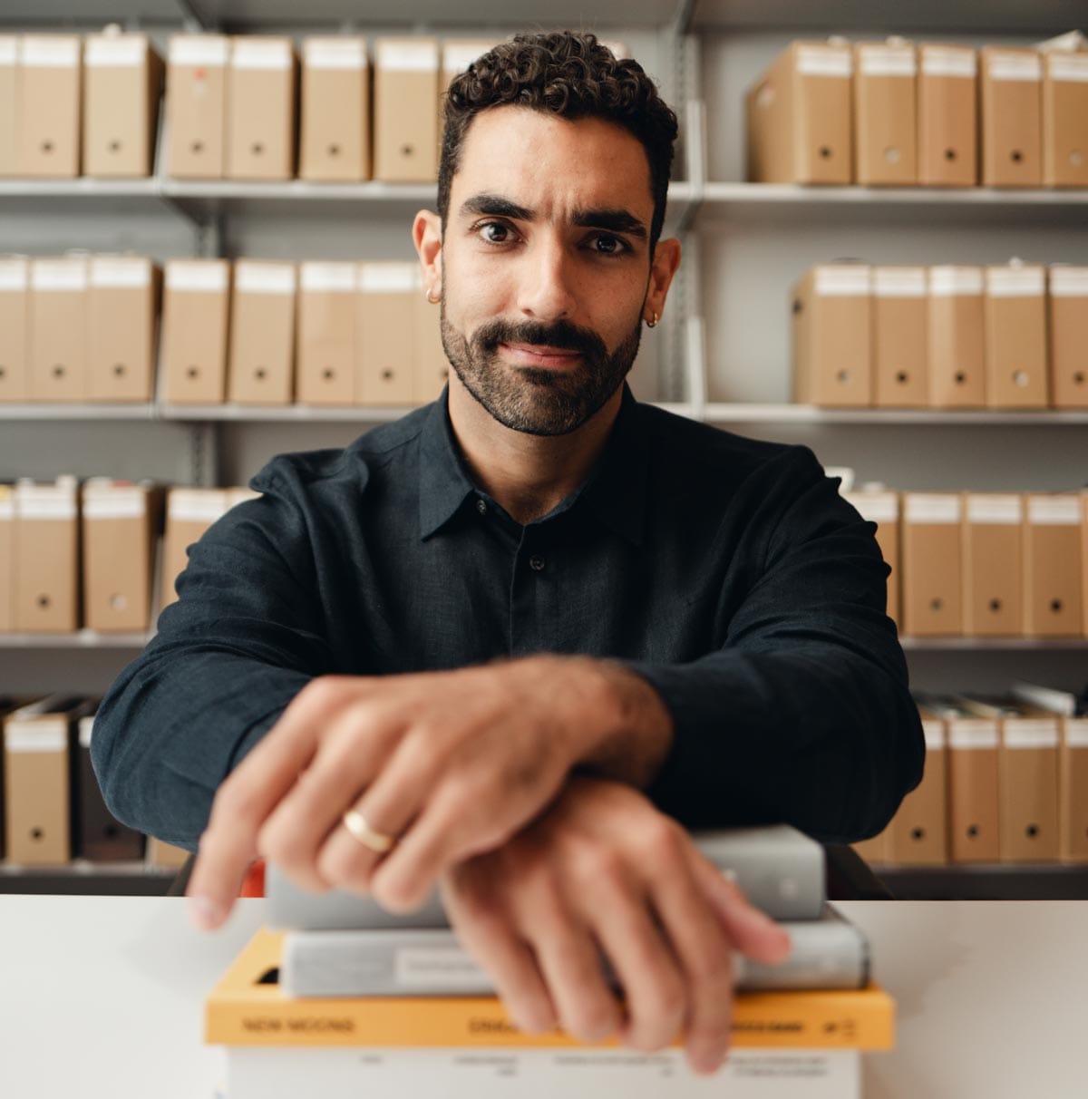 Bernardo sitting in an archival room with his hands-crossed over a pile of books.